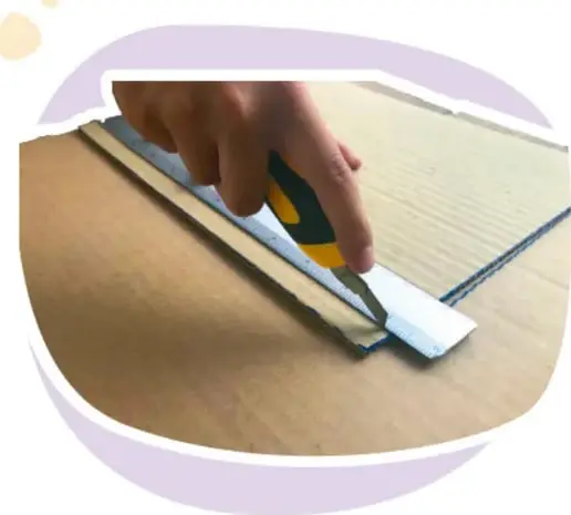 Mano de un adulto usando un cúter para hacer un corte superficial sobre un cartón, con una regla metálica como guía.