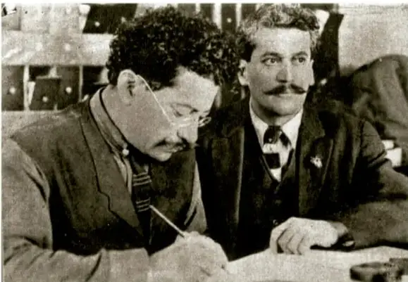 Fotografía en blanco y negro de dos hombres de la época: uno con lentes y bigote escribiendo, y otro con bigote mirando al frente, ambos con traje y corbata.