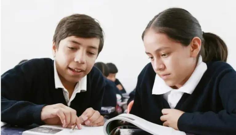 Fotografía de dos estudiantes de telesecundaria (un niño y una niña con uniforme azul marino y cuello blanco) revisando juntos un libro o revista sobre su pupitre.
