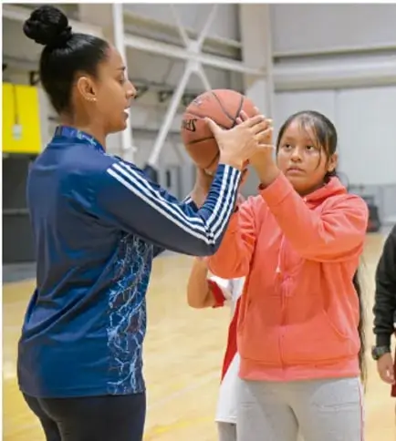 Fotografía de una entrenadora o instructora mostrando una pelota de baloncesto a una adolescente en un espacio deportivo cubierto, ilustrando la práctica de actividad física.