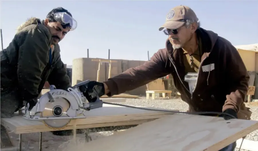 Fotografía de dos hombres trabajando con una sierra circular sobre una madera en un espacio abierto; uno de ellos usa lentes de protección y guantes, el otro lleva gorra y lentes oscuros. Al fondo se ven estructuras de madera y lonas.
