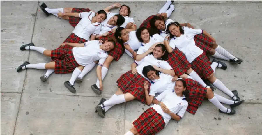 Fotografía cenital de un grupo de estudiantes con uniforme escolar (blusas blancas y faldas a cuadros rojas) tendidos en el piso formando una especie de flor o mandala radial, con los brazos entrelazados, transmitiendo unión y trabajo en equipo.