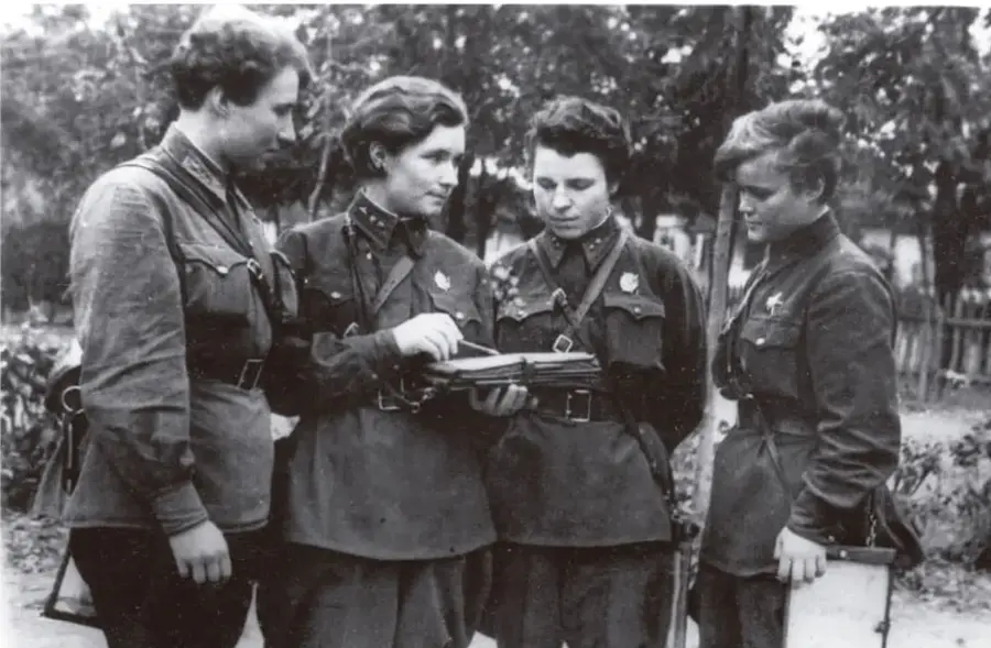 Fotografía en blanco y negro de cuatro mujeres con uniformes militares, reunidas alrededor de un mapa o documento, en un entorno al aire libre.