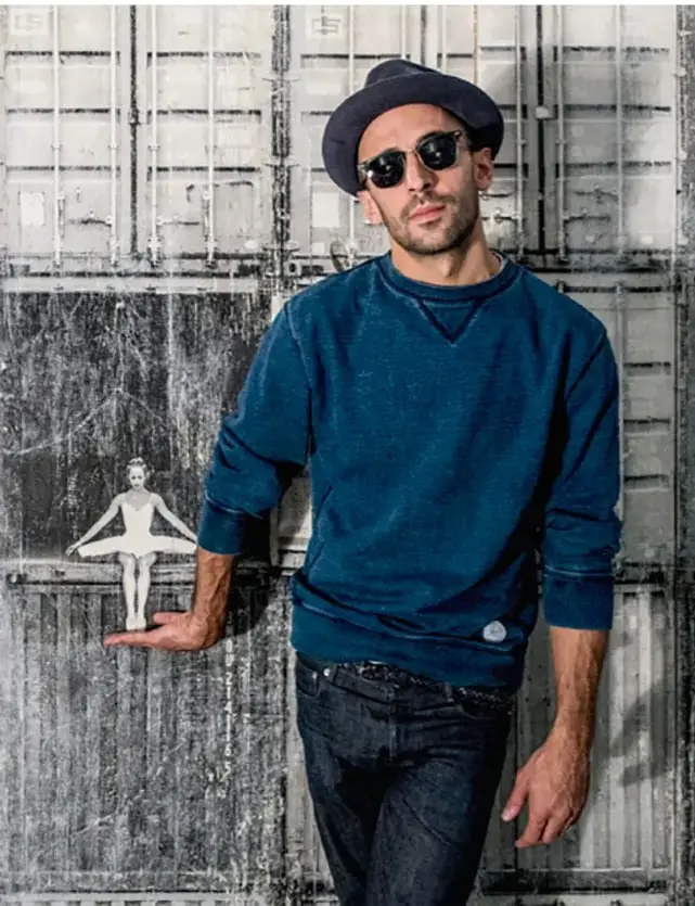 Fotografía del artista JR posando con sombrero y lentes, de pie frente a una pared gris con una pequeña figura de bailarina sostenida en su mano.