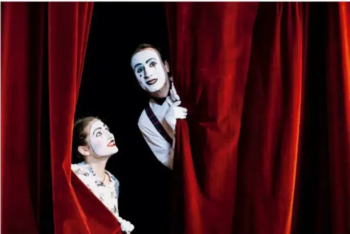 Dos personas con el rostro pintado de blanco (mimos), vestidas de blanco con detalles, asoman sonrientes entre cortinas rojas de terciopelo de un teatro.