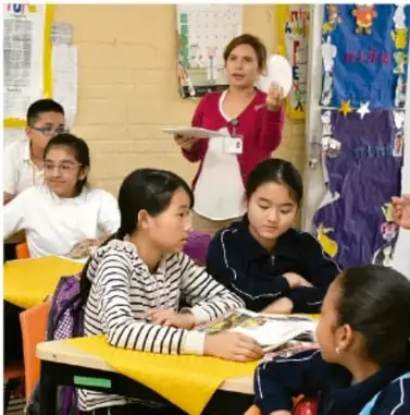 Fotografía de un salón de clases con una maestra de pie y varios estudiantes sentados en mesas, algunos con trípticos en las manos, durante una presentación.