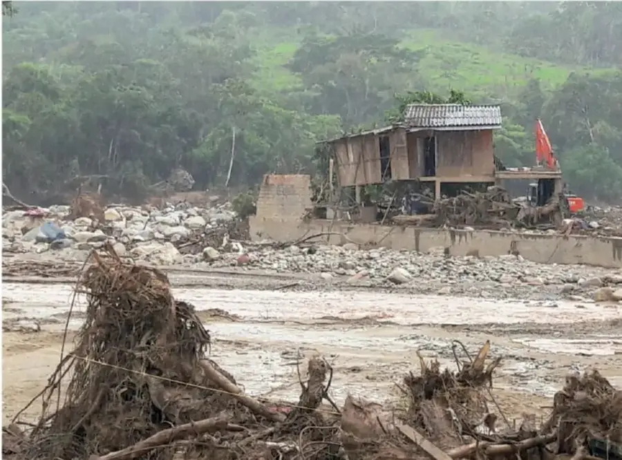 Fotografía de una comunidad afectada por un deslave o inundación: se observan lodo, piedras, troncos arrastrados, una casa de madera dañada y una excavadora naranja trabajando en la zona.