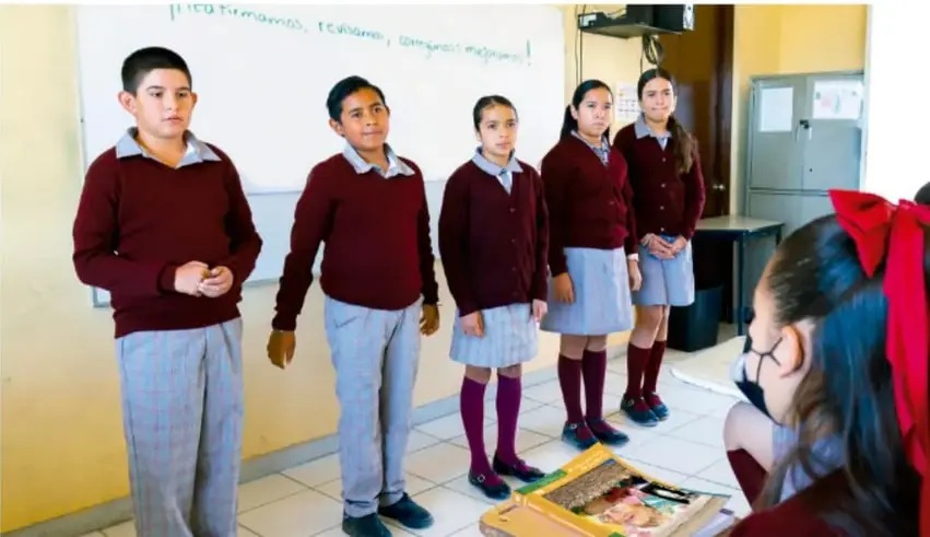 Cinco estudiantes de telesecundaria uniformados (suéter granate y pantalón/skirt gris a cuadros) de pie frente a la clase exponiendo; en primer plano una estudiante con moño rojo escucha. En el pizarrón se lee parcialmente '...lema, promovamos, compartamos ¡memorias mágicas!'.