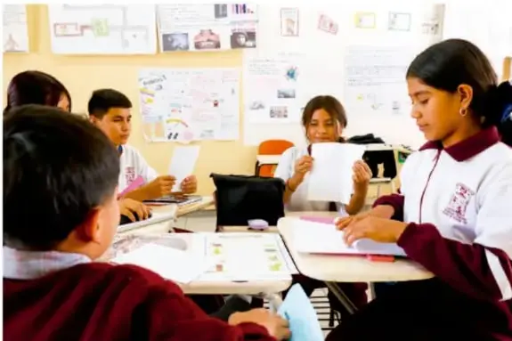 Fotografía de estudiantes de telesecundaria trabajando en equipo en el aula, revisando hojas y materiales sobre sus pupitres.