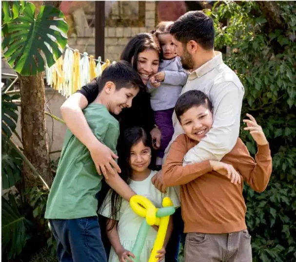 Fotografía de una familia (padres e hijos) abrazándose y sonriendo en un patio con plantas, ilustrando el sentido de pertenencia familiar.