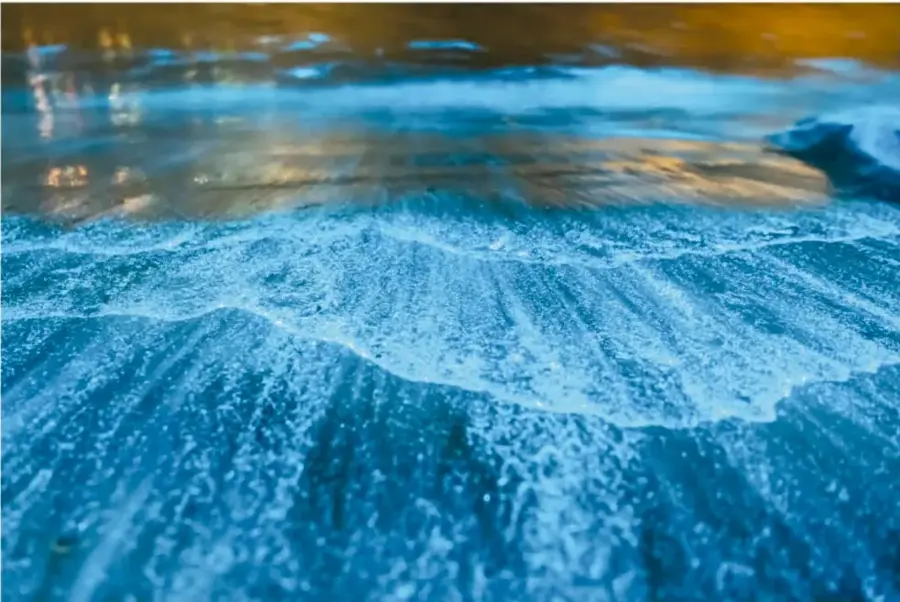 Fotografía en primer plano de olas del mar rompiendo en la orilla al atardecer, con espuma brillante color azul y reflejos dorados del sol.