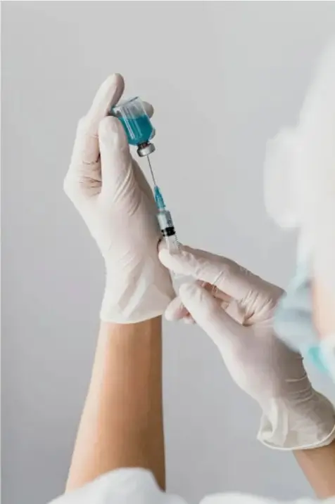 Fotografía de dos manos con guantes de látex: una sostiene un frasco o vial de vacuna y la otra aplica una jeringa, preparando la dosis de una vacuna.
