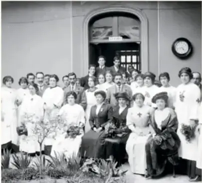 Fotografía antigua en blanco y negro de un grupo numeroso de personas, en su mayoría mujeres, frente a un edificio.