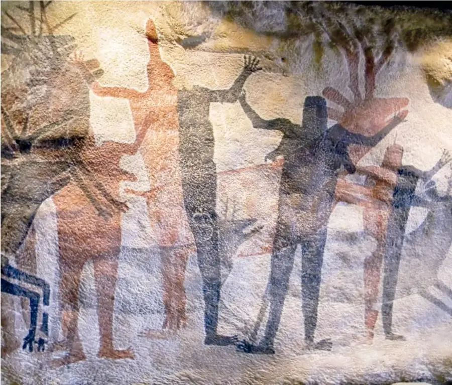 Gran mural rupestre sobre pared de roca clara: varias figuras humanas de cuerpo alargado y brazos levantados, pintadas en rojizo, ocre y azul oscuro, junto a ciervos y líneas de flechas.
