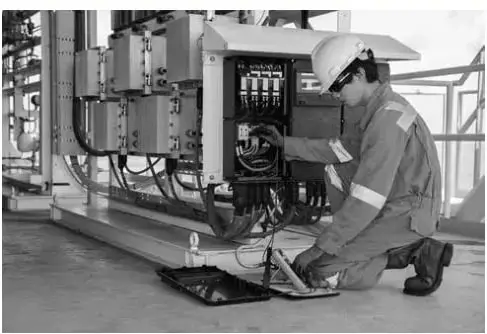 Fotografía en blanco y negro de un trabajador con casco y overol, arrodillado, manipulando una caja de conexiones eléctricas con cables; viste guantes de protección.