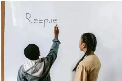 Niño escribiendo en un pizarrón la palabra 'Respue' mientras una niña lo observa.