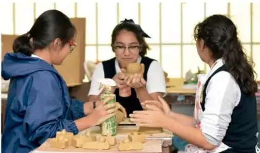 Tres estudiantes manipulando materiales y figuras de madera durante una actividad artística o de construcción.