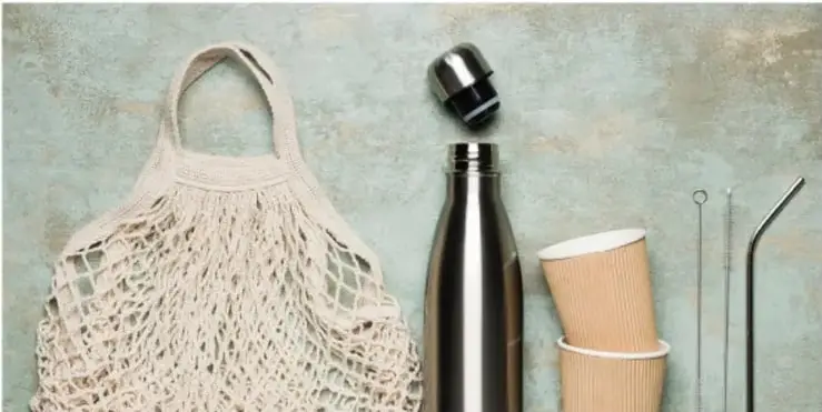 Objetos reutilizables: bolsa de red de tela, botella térmica de acero, vasos de cartón y popotes metálicos, sobre fondo verde.
