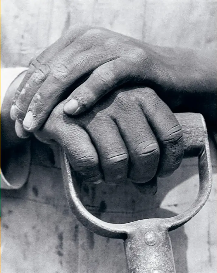 Fotografía en blanco y negro de dos manos superpuestas sujetando el mango de una herramienta de trabajo (como un pico o pala), representando el trabajo comunitario.