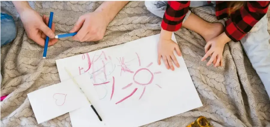 Fotografía de manos de niños y un adulto dibujando con plumones sobre una hoja blanca, sobre una manta tejida. Se observa un dibujo de un sol y una tarjeta con un corazón.