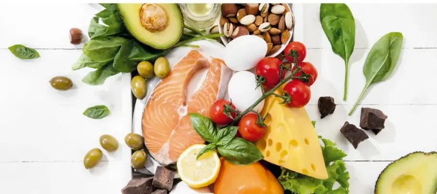 Fotografía de alimentos saludables: salmón, aguacate, espinacas, huevos, tomates, queso, frutos secos, chocolate oscuro, limón y aceitunas sobre mesa de madera blanca.