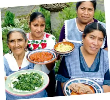 Fotografía de cuatro mujeres de distintas edades, con blusas bordadas, sosteniendo platillos tradicionales como quelites, mole y tamales; al fondo se ve vegetación.