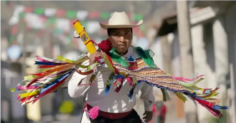 Fotografía de una persona con sombrero de ala ancha y camisa blanca sosteniendo un palo adornado con muchas cintas de colores (rojo, verde, amarillo, rosa, azul), probablemente durante una danza o celebración tradicional con banderines al fondo en una calle.