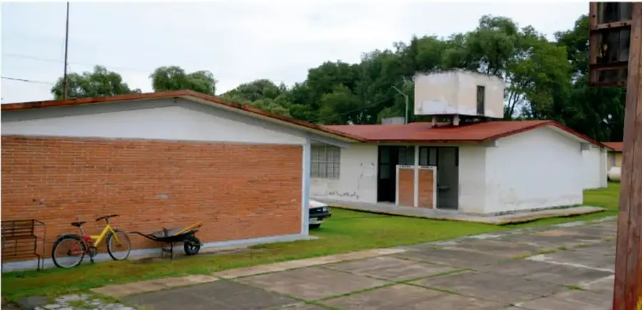 Fotografía de una escuela de telesecundaria: edificio de un piso con muro de ladrillo y muro blanco, techo rojo, patio pavimentado con césped, una bicicleta roja amarrada junto a una carretilla, árboles al fondo y un poste de madera a la derecha.