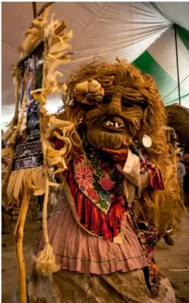 Fotografía de una figura disfrazada con máscara y vestimenta de fibras naturales, probablemente una tradición o danza folclórica mexicana, con adornos de colores y un estandarte; crédito: Sandra Angélica Martínez Cruz, INAH, CULTURA.