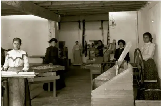 Fotografía en blanco y negro de una fábrica textil durante el Porfiriato. Se observan varias mujeres trabajando en máquinas industriales, con vestimenta de la época (blusas claras y faldas largas). Al fondo hay más trabajadoras y equipo industrial.