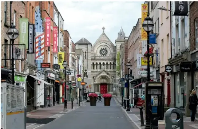 Calle peatonal de una ciudad de habla inglesa con tiendas, iglesia al fondo y transeúntes.