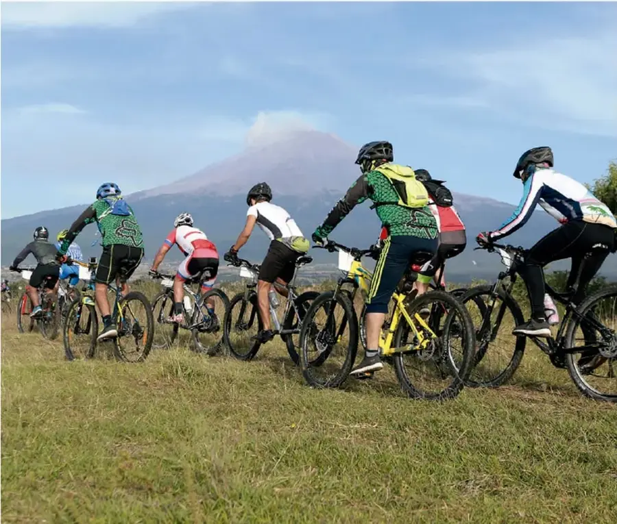 Grupo de ciclistas de montaña (entre 6 y 8) con casco y ropa deportiva, circulando por un camino de pastizal; al fondo se observa un volcán con columnas de vapor y cielo azul con nubes.