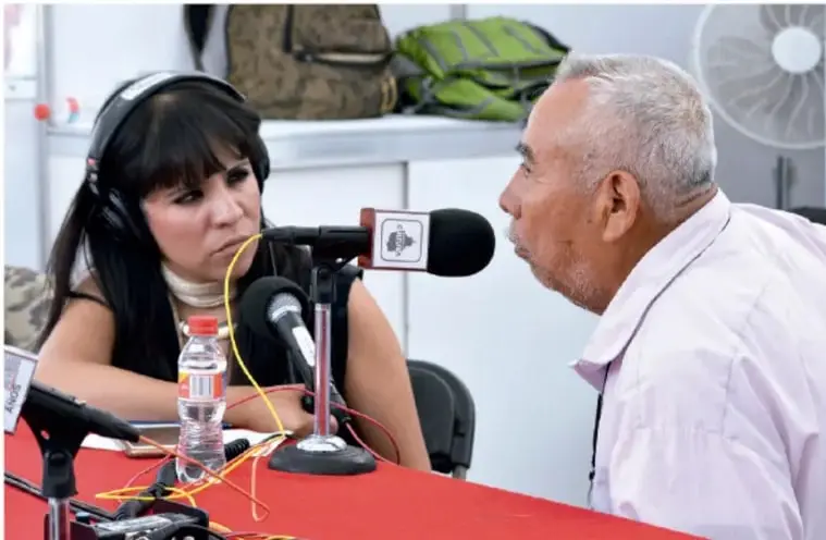 Fotografía de una entrevista en estudio de radio: una joven entrevistadora con audífonos frente al micrófono y un hombre mayor como entrevistado.