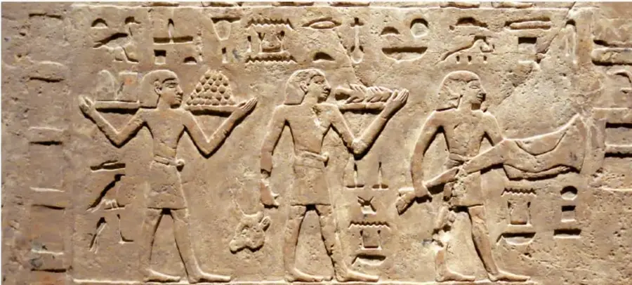 Relieve en piedra con figuras humanas que portan cestas y productos; a su alrededor hay signos grabados. Representa una escena de trabajo o entrega de productos en una civilización antigua.