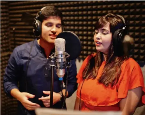 Fotografía de dos jóvenes (varón y mujer) con audífonos frente a un micrófono de estudio, grabando un programa.