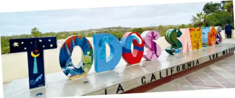 Letras monumentales de colores con la palabra TODOS SANTOS, cada una pintada con motivos artísticos, sobre una base con el texto BAJA CALIFORNIA SUR MÉXICO, al aire libre.