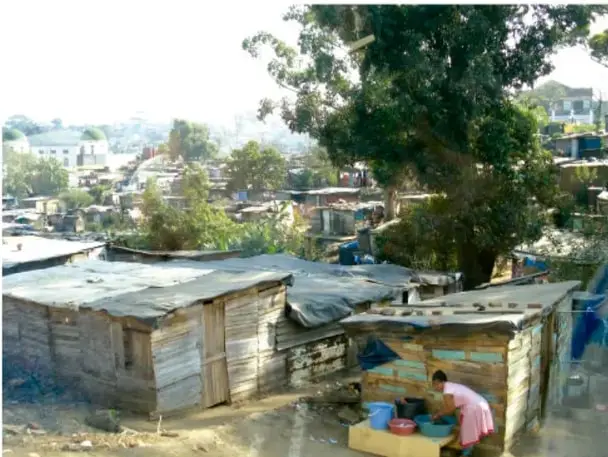 Fotografía de un asentamiento urbano marginal con viviendas de materiales precarios (láminas, madera), techos improvisados, árboles y una persona realizando actividades domésticas al aire libre, ilustrando las condiciones de pobreza descritas en el texto.