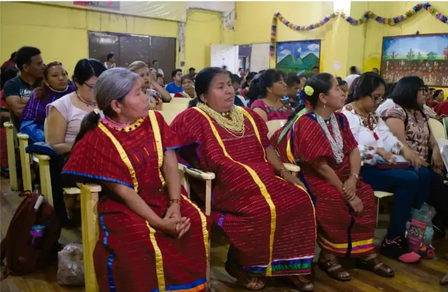 Fotografía de mujeres indígenas (pueblo maya) con huipiles rojos, fajas y collares, sentadas en bancas dentro de un salón decorado con guirnaldas, participando en un evento o asamblea comunitaria.