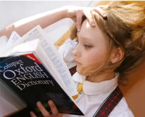 Fotografía de una estudiante con uniforme escolar leyendo detenidamente un Compact Oxford English Dictionary, apoyada la cabeza en la mano, ilustrando el estudio del idioma inglés.