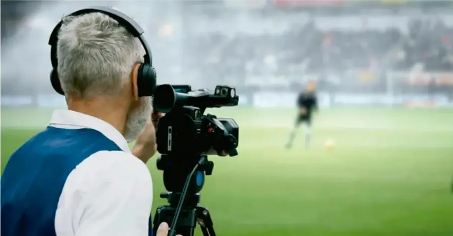 Fotografía de un camarógrafo de espaldas, con audífonos y chaleco azul, filmando un partido de fútbol en un estadio con un jugador difuso de fondo.