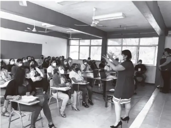 Fotografía en blanco y negro de una sesión de capacitación: una expositora de pie, con cubrebocas, gesticula frente a un grupo de participantes adultos sentados con cubrebocas en un salón con pizarra, ventanas y ventiladores de techo.