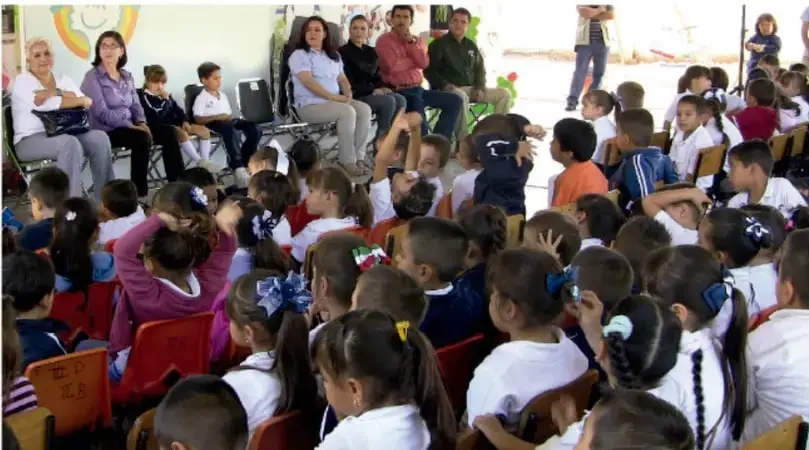 Evento comunitario en el patio escolar: adultos sentados y muchos niños de uniforme participando.