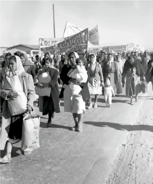 Fotografía en blanco y negro de una multitud de personas, principalmente mujeres con niños, caminando en una marcha. Llevan mantas y pancartas, una de ellas dice 'LOS MINEROS...'. Se observa un ambiente de protesta y desplazamiento.