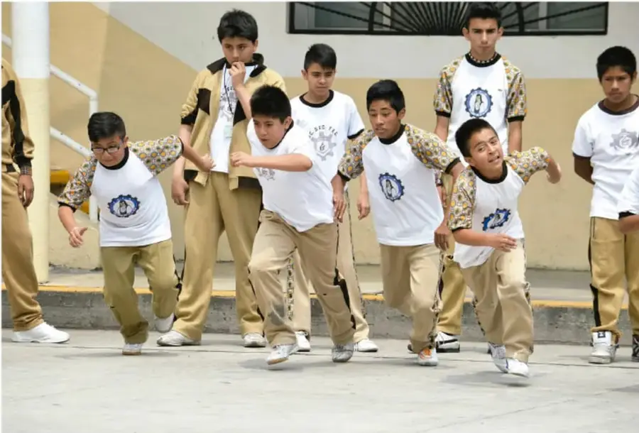 Grupo de adolescentes con uniforme escolar (camiseta blanca y pantalón beis) realizando actividad física en el patio de su escuela, algunos corriendo y otros observando.