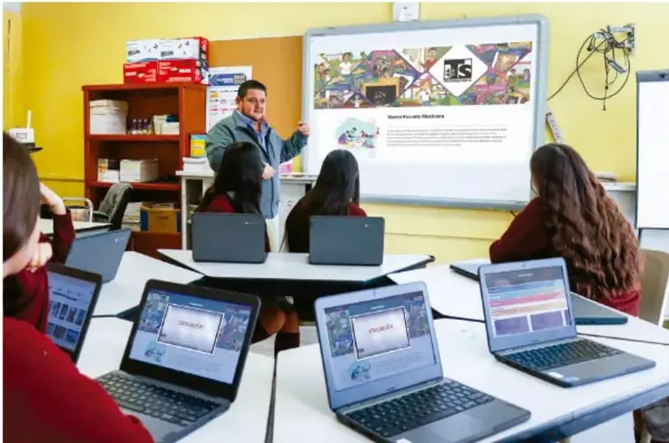 Aula de Telesecundaria: un docente frente a una pantalla interactiva presentando un sitio web, con estudiantes sentados en mesas trabajando en computadoras portátiles.