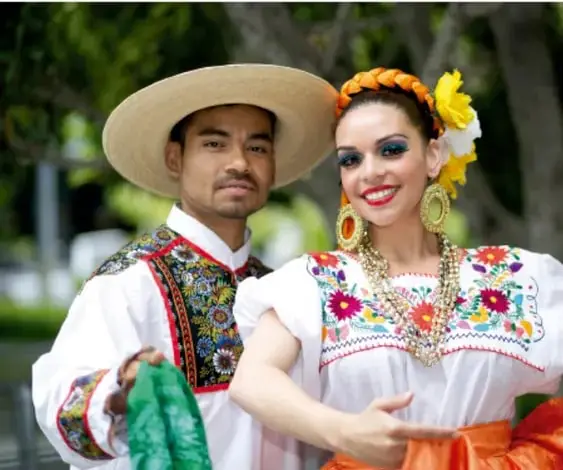 Fotografía de una pareja vestida con trajes típicos mexicanos: el hombre con sombrero de paja y camisa bordada, la mujer con huipil bordado, flores en el cabello y abundante joyería, representando la diversidad cultural y lingüística de México.