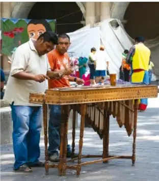 Fotografía de músicos que tocan la marimba de madera en una explanada pública al aire libre, con carpas y público al fondo.