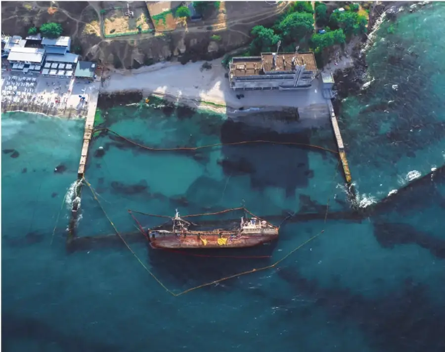 Fotografía aérea de un buque varado o encallado en aguas costeras, rodeado por una barrera de contención flotante (booms) que intenta acotar una mancha oscura de contaminante en el agua; al fondo se observan edificios y vegetación en la costa.
