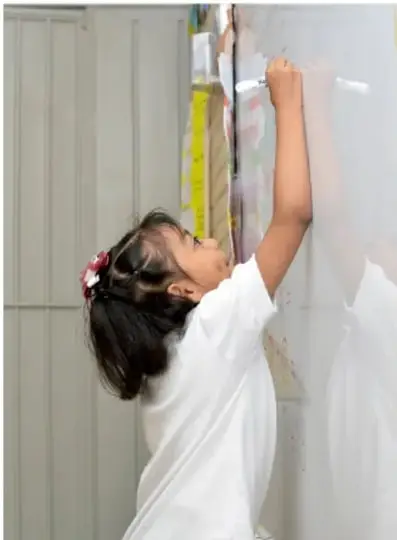 Niña de espaldas escribiendo o pegando carteles en un pizarrón o muro, como parte de la organización de actividades escolares.