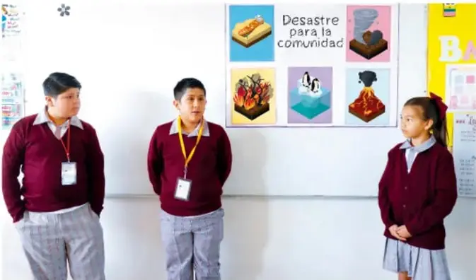 Fotografía de tres estudiantes de Telesecundaria con uniforme (suéter granate con gafete y pantalón/sketch gris) de pie frente a un salón; detrás hay un cartel del proyecto con el título "Desastre para la comunidad" y varios imágenes de desastres (incendio, tornado, inundación, volcán) y otros carteles escolares.